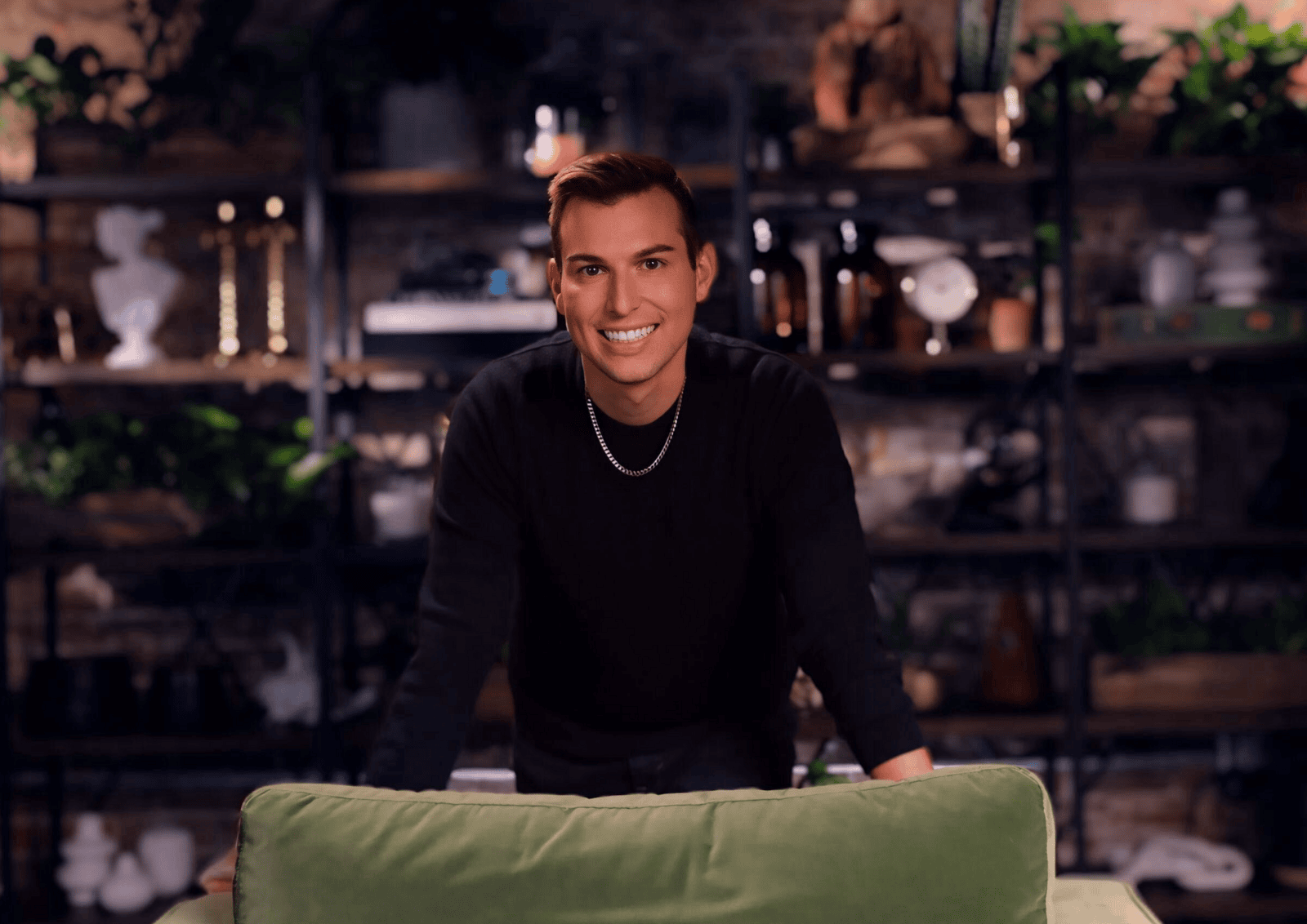 Matt Fraser, a man with short brown hair, wearing a black shirt and a gold chain necklace, stands behind a green couch in a room with shelves and plants in the background.